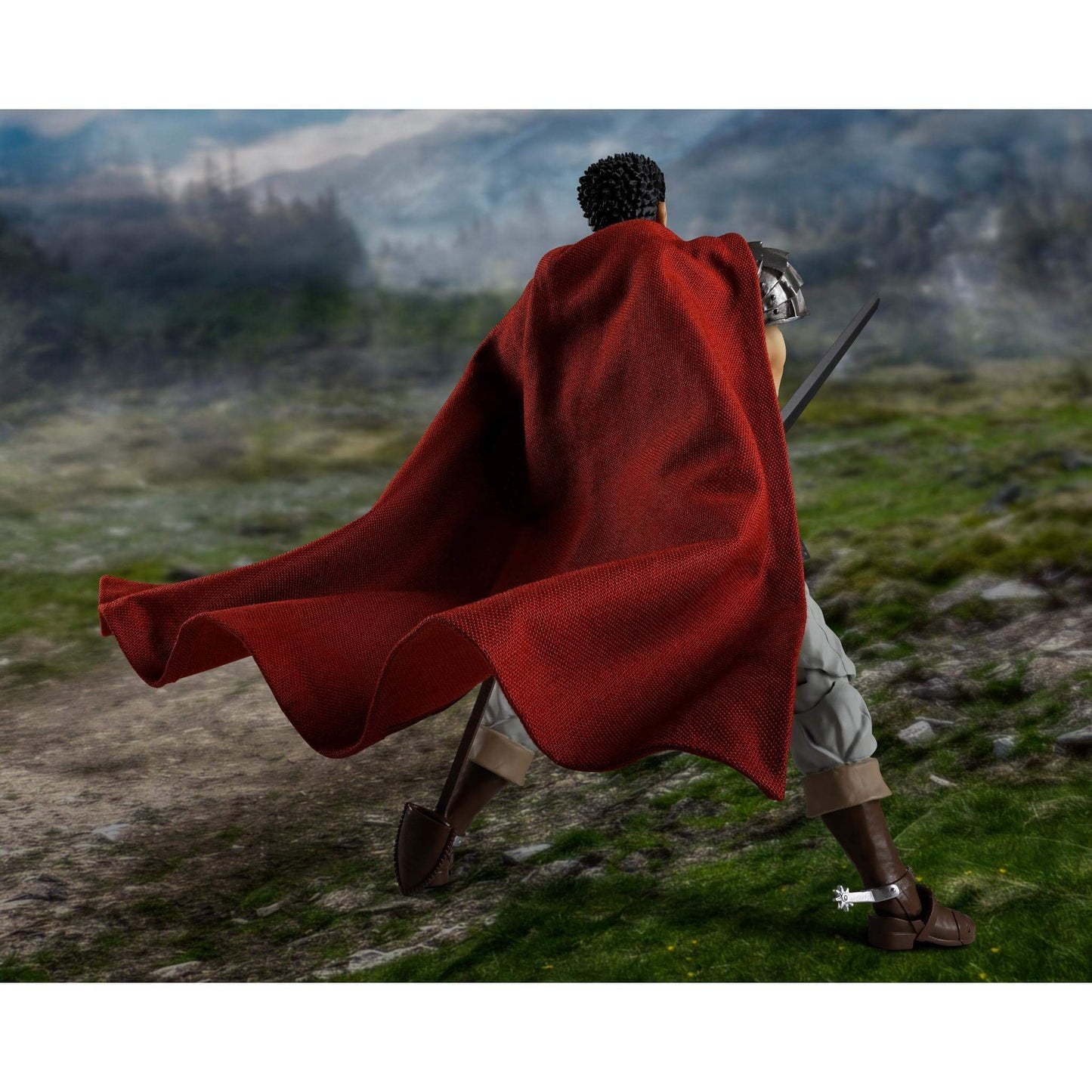 Berserk S.H.Figuarts Action figure of Guts in a red cape and sandals walking on a grassy hill with mountains in the background, back facing